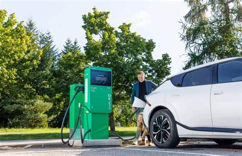 Tematinė nuotrauka, vaizduojanti elektromobilį įkrovimo stotelėje, o fone matoma saulės energijos jėgainė