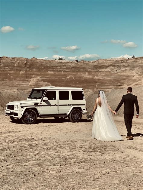 Jaunavedžiai pozuoja prie balto „Mercedes-Benz G“ klasės automobilio vestuvių fotosesijos metu